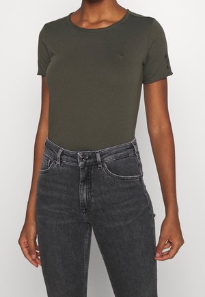 Femme portant un t-shirt ajusté vert olive à manches courtes et un jean noir délavé taille haute sur un fond uni.