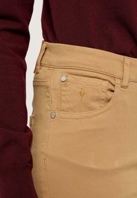 Pantalones color marrón con una textura suave y detalles en los bolsillos frontales. Presenta herrajes plateados y sutiles acentos de costura. Combinados con una blusa de color rojo oscuro.