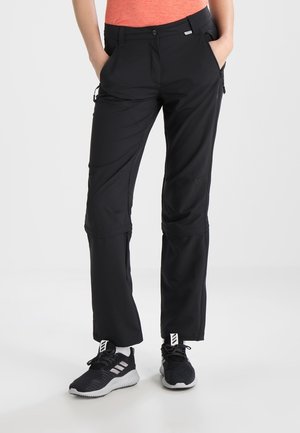 Frau trägt schwarze Wanderhose mit Reißverschluss und Seitentaschen sowie schwarze Sportschuhe, steht mit den Händen in den Taschen.