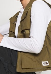 Person som bär en olivgrön North Face-väst över en vit långärmad tröja, i mörka byxor, med armarna böjda och vilande på knäna.