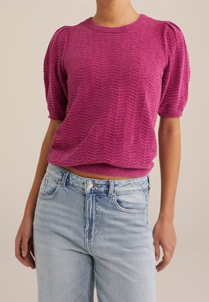 Femme portant un pull à manches courtes texturé rose vif avec des épaules bouffantes, rentré dans un jean en denim bleu clair.
