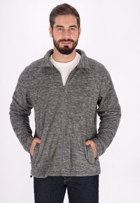 Giacca in pile grigia con zip, dalla finitura testurizzata, colletto rialzato e tasche laterali, indossata sopra una camicia bianca. Tessuto morbido e caldo.