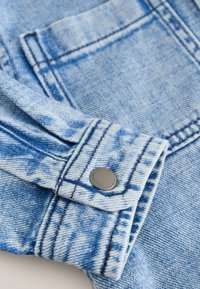 Hellblauer Jeansjackenärmel mit sichtbarer Naht und einem Metall-Druckknopfverschluss nahe dem Bündchen.
