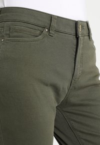Pantalones de algodón verde oliva con un ajuste Slim, diseño de cinco bolsillos y herrajes de metal. La tela tiene una textura suave con costuras visibles.