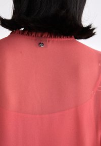 Volánový výstřih z průsvitné červené tkaniny, s černým logem a knoflíkovým detailem na zadní straně. Hladká textury s lehkým vzhledem.