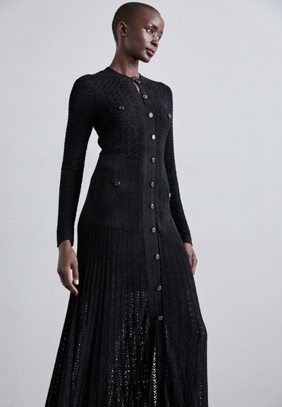 Sort strikket kjole med lange ærmer, knapfront, teksturerede mønstre og to brystlommer, samt et plisseret skørtdesign.