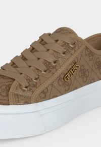 Καφέ αθλητικό παπούτσι Guess με σχέδιο, μπεζ κορδόνια, χρυσά οπές και λευκή λαστιχένια σόλα.