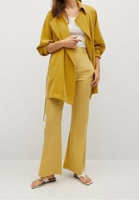 Tenue jaune moutarde avec une chemise ample à manches longues et un pantalon évasé, associée à des sandales plates beiges ; design simple, texture lisse.
