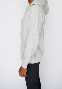 Sudadera gris con cremallera frontal, mangas largas y capucha. Cuenta con un bolsillo tipo canguro y puños acanalados, usada sobre pantalones negros.