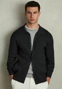 Cardigan negru cu nervuri, cu închidere pe bază de nasturi, purtat peste un tricou gri deschis cu guler rotund, asociat cu pantaloni crem. Design simplu și siluetă ajustată.