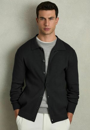 Cardigan negru cu nervuri, cu închidere pe bază de nasturi, purtat peste un tricou gri deschis cu guler rotund, asociat cu pantaloni crem. Design simplu și siluetă ajustată.
