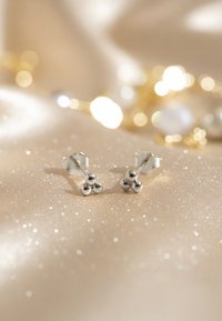 Silversked örhängen med tre sfäriska detaljer, mot en glittrande beige bakgrund. Slät textur med en polerad finish.