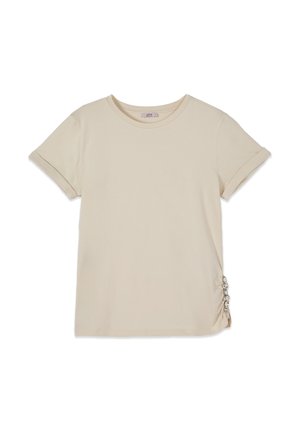 Beige t-shirt met korte mouwen, opgerolde zomen en decoratieve zilveren ringen die de stof aan de rechterbenedenkant samenbrengen.
