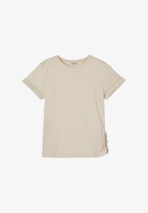 Beżowy t-shirt z krótkim rękawem, z podwiniętymi mankietami i dekoracyjnymi srebrnymi pierścieniami zbierającymi materiał po prawej dolnej stronie.