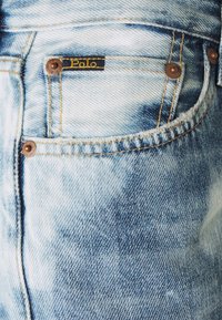 Světle modré džíny z denimu se zesvětlenými místy, s černou nášivkou s nápisem "Polo" a bronzovými kování na kapsách.