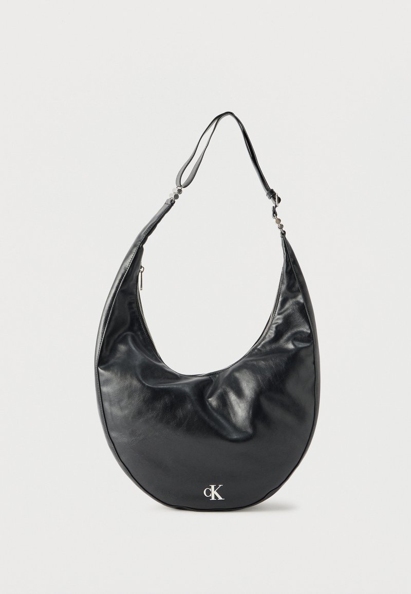 Μαύρη δερμάτινη τσάντα ώμου σε σχήμα ημισελήνου με ρυθμιζόμενο λουρί και ασημένιο λογότυπο "cK" στο μπροστινό μέρος, με φόντο λευκό.