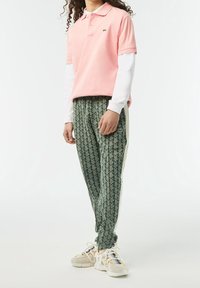 Polo rose avec un col replié, associé à un pantalon de survêtement vert à motifs et des baskets blanches texturées. Les accents incluent un logo sur la chemise.