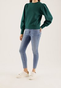 Groene katoenen pullovertrui met pofmouwen, gecombineerd met lichtblauwe skinny jeans en witte sneakers met gouden accenten.