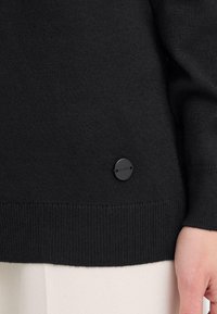 Schwarzer, gestrickter Pullover mit einem gerippten Saum und einem runden schwarzen Logo-Patch an der Seite. Glatte Textur; figurbetonte Silhouette.