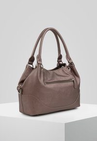 Brązowa skórzana torebka na podwójnych uchwytach, z przednią kieszenią na zamek błyskawiczny i srebrnymi metalowymi pierścieniami, stojąca na białym piedestale na szarym tle.