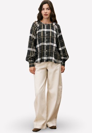 Femme portant une blouse à carreaux noire et beige avec des manches bouffantes et un pantalon crème à jambes larges, se tenant contre un fond clair.