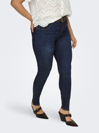 Nainen, joka pukeutunut tummiin kapeisiin farkkuihin, valkoiseen silmukkahuiviseen paitaan ja tummiin ruskeisiin, kärjestä teräväkärkisiin kiilakorkokenkiin, joissa on solkikiinnitteiset remmit, seisoo käsi taskussa.