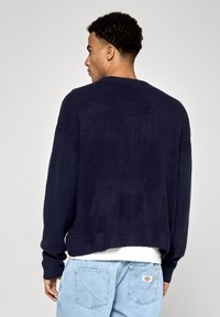 Námorně modrý pletený svetr s uvolněným střihem, žebrovaným výstřihem a manžetami, širokými rukávy a hladkou texturou, nošený přes bílou košili.
