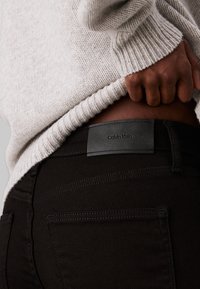 Jeans de mezclilla negra con una textura suave y costuras sutiles; cuentan con una etiqueta de cuero negro de Calvin Klein en la cintura.