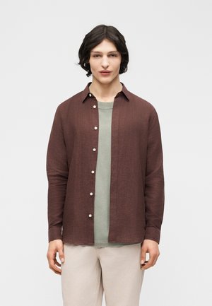 Giovane con capelli castano scuro di media lunghezza indossa una camicia marrone aperta con bottoni sopra una maglietta verde chiaro e pantaloni beige chiaro.