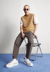 Knit tan vest over a white t-shirt, gray trousers, and white high-top sneakers. Individual with sunglasses seated on a chrome chair. Blue background.