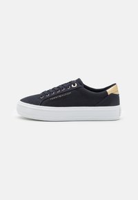 Nízké modré tenisky s bílou podrážkou, zlatým akcentem na patě a logem "Tommy Hilfiger" na boku, zobrazené z vnější strany.