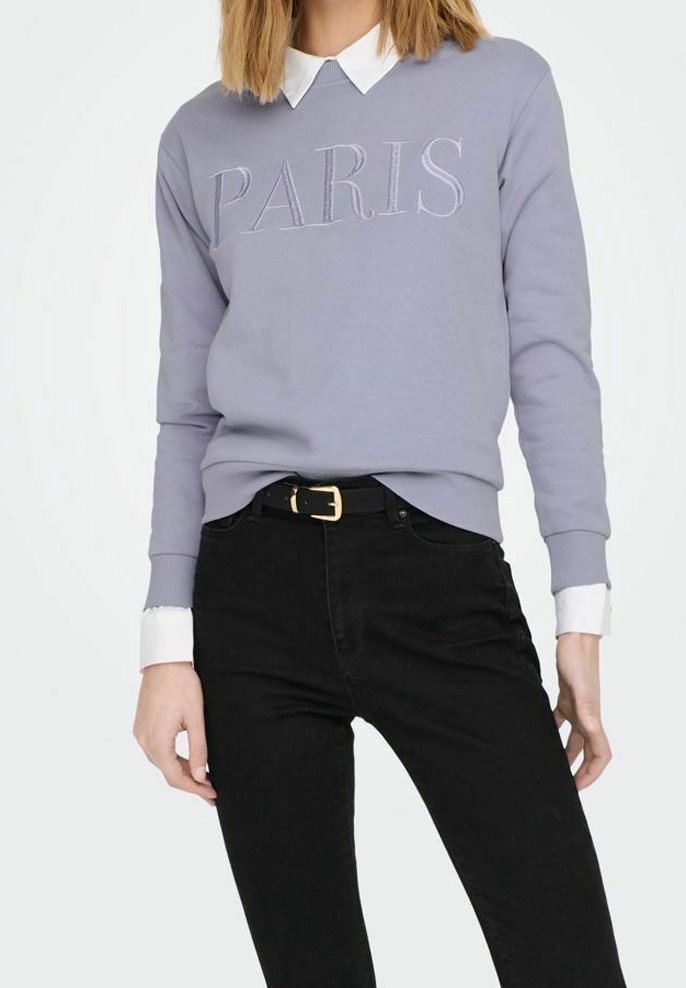 Felpa lavanda con "PARIS" ricamato in bianco, abbinata a jeans neri e una camicia bianca con colletto sotto. Tessuto morbido e casual.