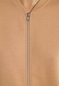 Prenda con cremallera en color beige, con una cremallera plateada visible, que muestra una textura de tela suave y un cuello redondo.