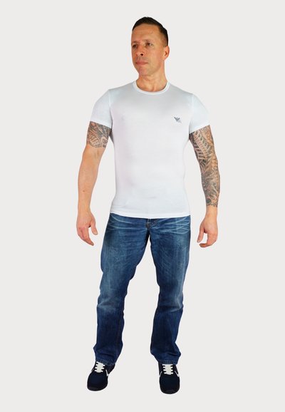 Weißes, eng anliegendes T-Shirt mit kurzen Ärmeln, minimalem Branding auf der Brust, Jeans mit ausgewaschenem Denim und schwarze Sneakers mit weißen Akzenten.