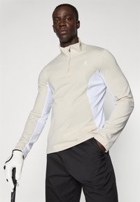 Béžový golfový top s dlhým rukávom a golierom na polovičný zips, biele bočné panely; vyrobený z hladkej tkaniny. Model drží golfovú palicu v rukavici.