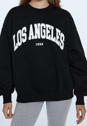 Osoba ubrana w czarną bluzę z napisem "LOS ANGELES 1990" w białym kolorze oraz jasnoszare legginsy, stojąca na tle jednolitego tła.