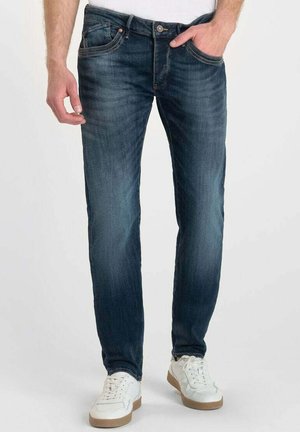 Jeans de denim azul con un acabado desgastado, diseño de pierna recta, estilo de cinco bolsillos y herrajes en tono cobre. Combinados con zapatillas blancas.