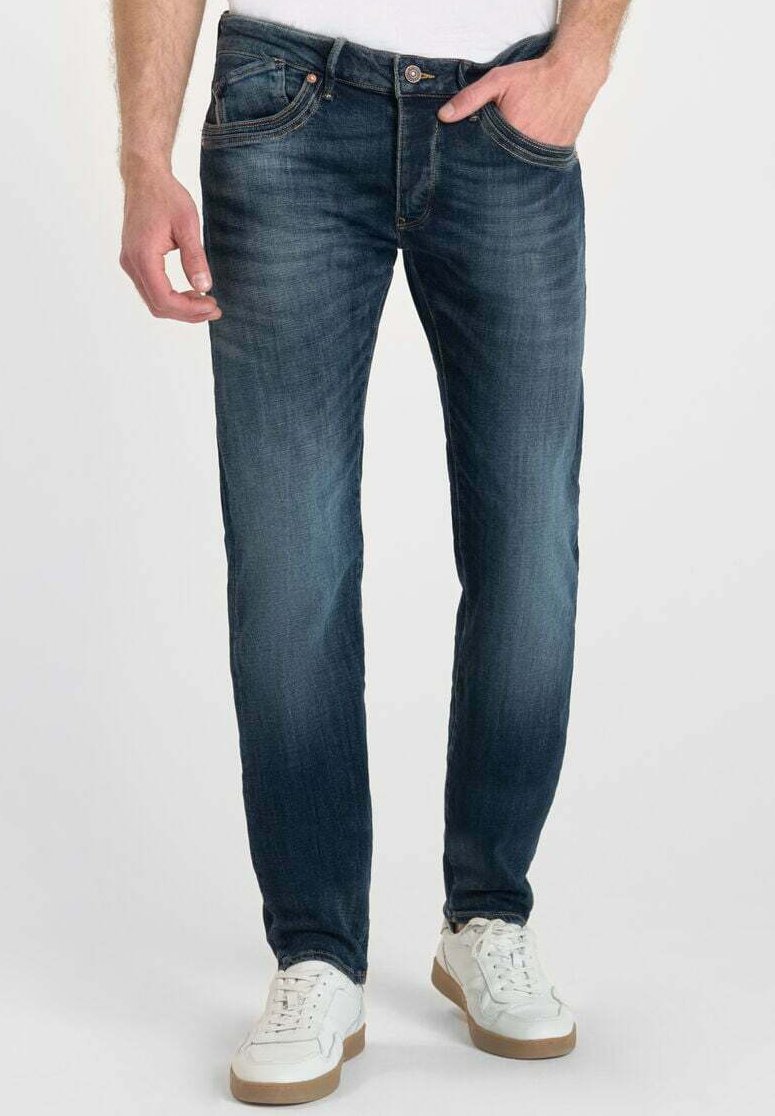 Jeans de denim azul con un acabado desgastado, diseño de pierna recta, estilo de cinco bolsillos y herrajes en tono cobre. Combinados con zapatillas blancas.