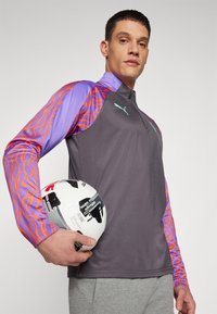 Szürke sportdzseki lila és narancssárga hullámos mintázatú ujjal, cipzáras nyakkal és logóval; egy fekete-fehér mintázatú focilabdát tart.
