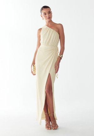 Žena v krémových jednom ramennom šatách s nariasením a bočným rozparkom, so zlatými doplnkami, držiaca zlaté textúrované psaníčko, stojaca pred bielym pozadím.