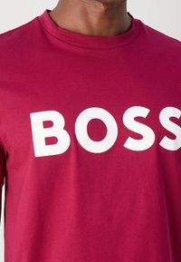 Homme portant une chemise rouge avec de grandes lettres blanches formant le mot "BOSS" sur la poitrine, cadré au niveau du cou et des épaules.