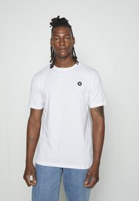 Vit bomulls t-shirt med rund halsringning och korta ärmar, prydd med en liten svart logotyp på bröstet. Matchad med blå denimjeans.