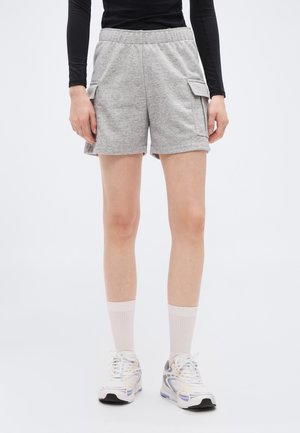 Persona valkā pelēkas cargo šortus, baltas sporta zeķes un baltus sporta apavus, stāv uz gaiša, vienveidīga fona.