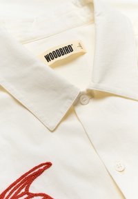 Chemise col blanc cassé avec des boutons blancs, ornée d'un détail brodé rouge et d'une étiquette de marque WOODBIRD beige à l'intérieur du col.