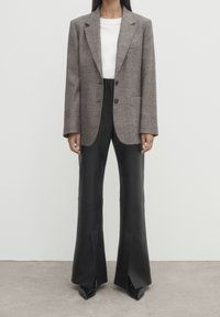 Blazer oversize marrone a quadri con due bottoni, abbinato a pantaloni neri a vita alta con fondo svasato e tacchi neri a punta. Texture lisce in tutto.