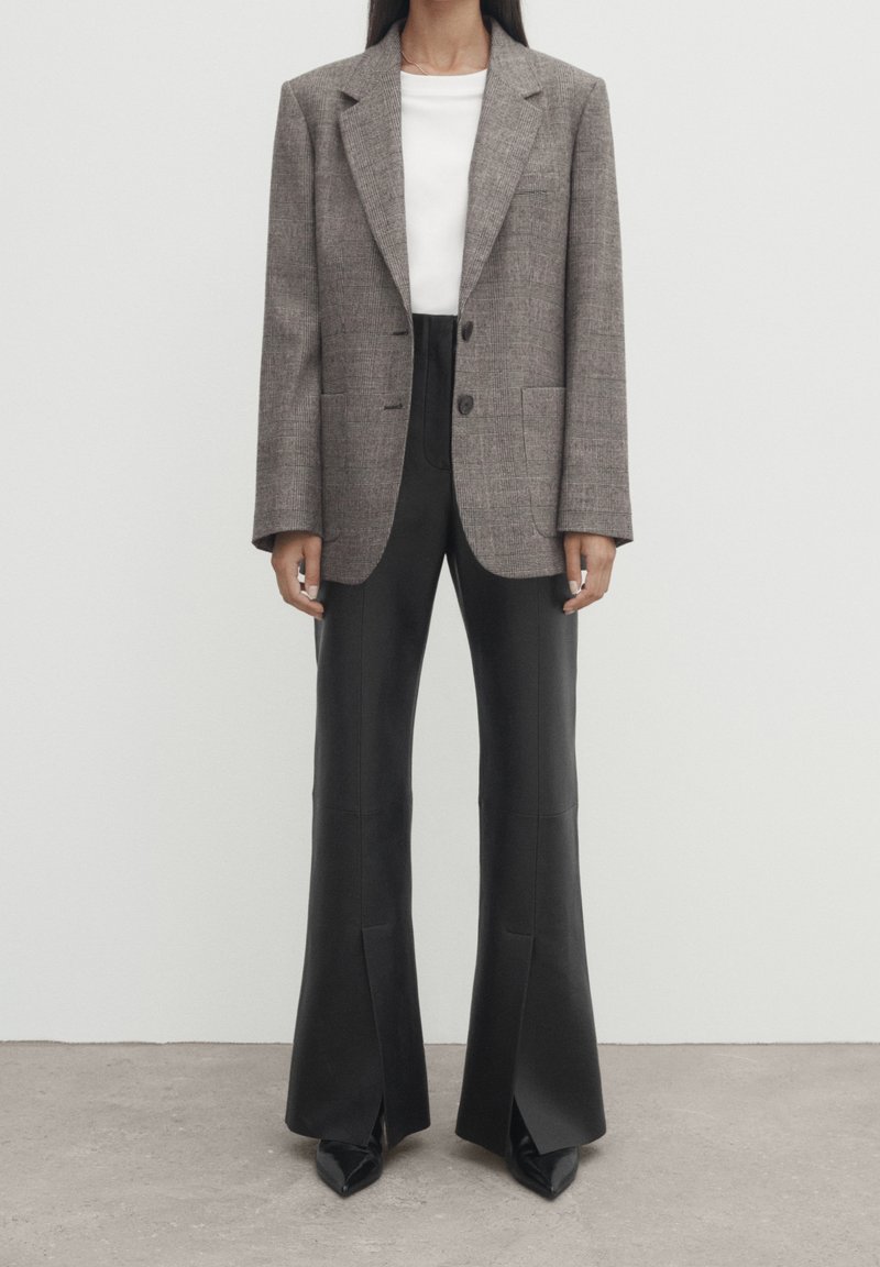 Blazer oversize marrone a quadri con due bottoni, abbinato a pantaloni neri a vita alta con fondo svasato e tacchi neri a punta. Texture lisce in tutto.