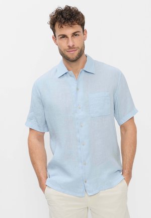 Mann mit kurzen lockigen Haaren, trägt ein hellblaues kurzärmeliges Hemd mit Knöpfen und beige Hose, Hände in den Taschen, neutraler Hintergrund.