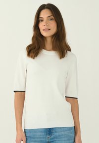 Kortärmad vit stickad tröja med rund hals, med svart kant vid ärmsluten och en texturerad yta. Bärs med denimjeans.