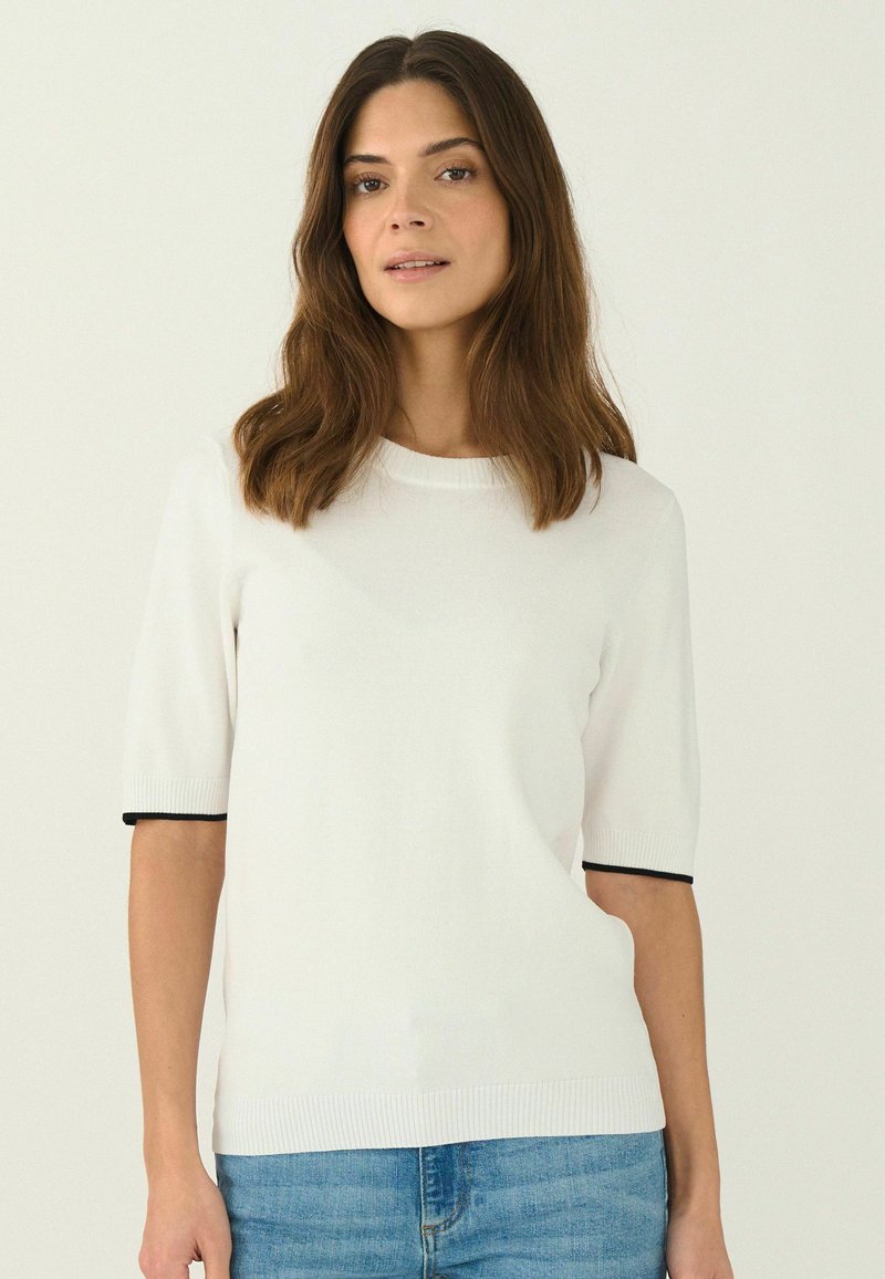 Kortärmad vit stickad tröja med rund hals, med svart kant vid ärmsluten och en texturerad yta. Bärs med denimjeans.