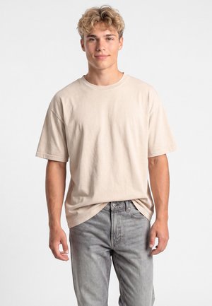 Giovane uomo con capelli biondi ricci che indossa una maglietta beige oversize e jeans grigi, in piedi davanti a uno sfondo bianco semplice.